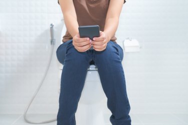 Woman sitting on toilet and using smartphone, showing the concept of legs falling asleep on the toilet