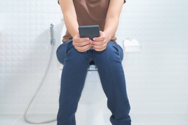 a close up of a person wearing jeans and a brown shirt sitting on the toilet and using their smartphone, showing the concept of legs falling asleep on the toilet, in an all-white bathroom