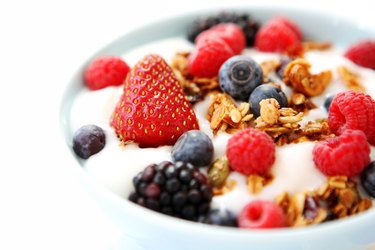 a close up of a white bowl of yogurt with fresh strawberries, blueberries, raspberries and blackberries and granola on a white background