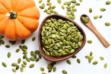 an overhead photo of pumpkin seeds in a wooden bowl with a small pumpkin on the side and a wooden spoon on a white background