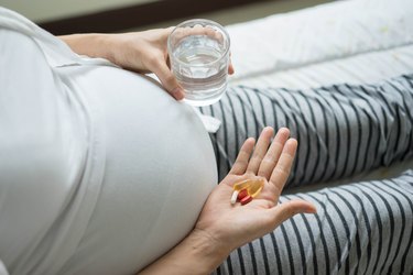 a close up photo of a pregnant person wearing a gray shirt and striped leggings holding pills in one hand with a glass of water in the other