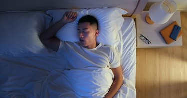 an overhead photo of a person wearing a white t-shirt sleeping in bed next to a night table with a lamp