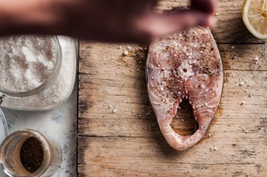 Seasoning a slice of Chub Mackerel fish steak with lemon, salt and ground pepper on a rustic cutting board