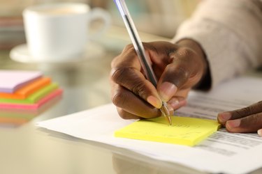 A person's hand writing on a sticky note on a desk