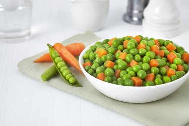 White bowl of cooked frozen peas and cubed carrots on white tabletop.