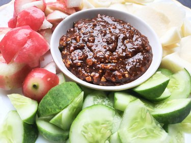Small bowl of rujak or rojak sauce on a white plate with assorted fruits and vegetables.