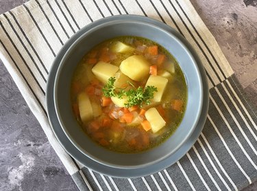 an overhead photo of a blue bowl of vegetable soup with potatoes, carrots, celery and parsley. on a striped linen napkin