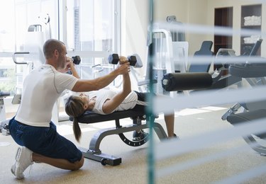 A personal trainer kneels behind an athlete on a bench and does a dumbbell chest press at a gym