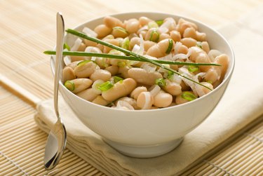 A white bowl filled with magnesium- and potassium-rich cooked cannellini beans and herbs on a tan tablecloth.