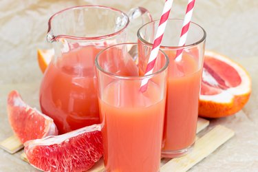 two glasses of red grapefruit juice with red and white striped straws next to a glass pitcher of grapefruit juice and slices of fruit on a light wooden background
