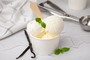Close-up of carbohydrate-rich ice cream or frozen yogurt with mint and vanilla pods on white textured table.