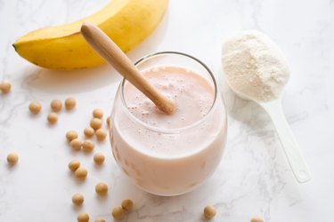 A close up of a protein shake in a glass on a white table with a banana and protein powder around it.