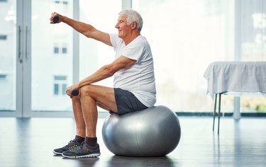 an older adult wearing a white t-shirt and black shorts sits on a silver stability ball holding a small dumbbell out in front of their face