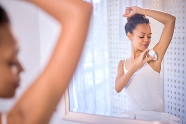 Person in a white tank top standing in front of a mirror in the bathroom holding a stick of deodorant and looking down at their armpit while applying it.