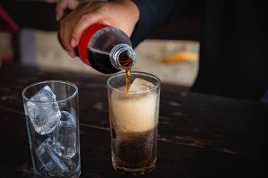 close up of a hand holding a cold cola bottle pouring it into a glass next to another glass of ice