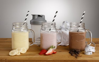 Banana, Strawberry and Chocolate milkshakes in Mason Jar glasses, with paper straws, ingredients and shaker on a wooden bench top