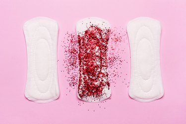 three menstrual pads, one with heavy red glitter, on a light pink background, to represent reproductive health symptoms