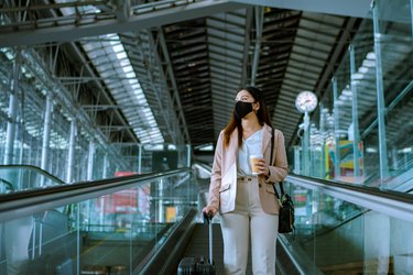 Asian businesswoman traveler with face mask on the move using escalator in airport.