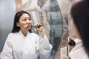 adult wearing a white bathrobe brushes their teeth with fluoride toothpaste in a hotel bathroom in front of a mirror