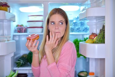 Woman with sugar cravings at night, sneaking a doughnut from the refrigerator.