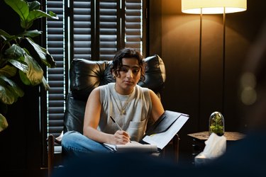 a transmasculine person sits in a leather chair in a gray tank and jeans writing in a notebook