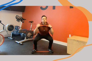 man with dark red shirt and black pants using a red resistance band under his feet, held in his hands and across his chest, doing a front squat for a leg and shoulder workout