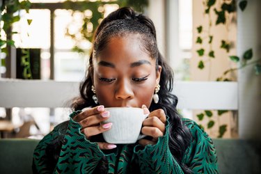 a young adult with a high ponytail takes a sip of coffee from a white mug in a cafe with plants in the background