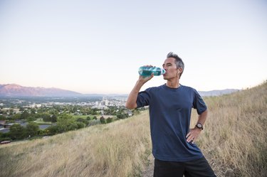 a middle aged adult drinks a healthy flavored water from a water bottle while hiking outside with mountains in the background