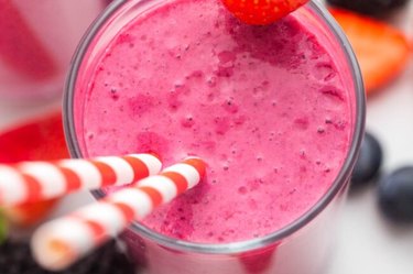 an overhead photo of a pink berry smoothie in a glass with two red and white striped straws and a strawberry on the rim