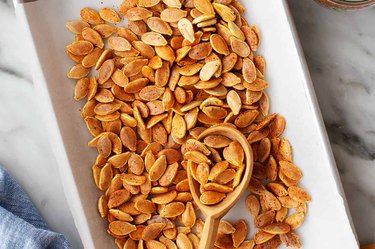 an overhead photo of a slim white platter of roasted, spiced pumpkin seeds with a wooden spoon holding some seeds