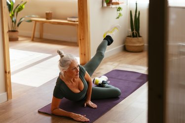 older person with gray hair in a bun does pilates side kicks for knee pain on a purple mat in a home