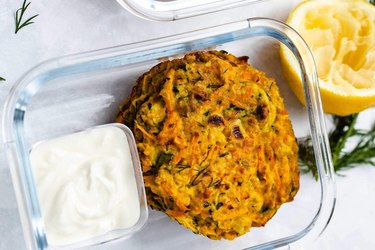 an overhead photo of a glass meal prep container of Zucchini Carrot Fritters next to a squeezed half of a lemon and dipping sauce