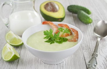Creamy Avocado Soup With Shrimp in a white bowl next to raw avocado, milk, and lime on a gray wooden table.