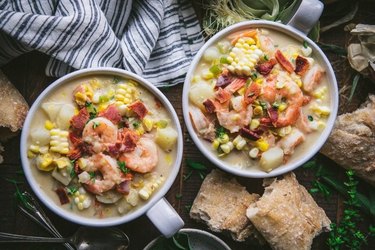 Shrimp Soup With Corn and Bacon in two white bowls next to raw ingredients on a table.