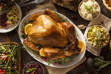 an overhead photo of a pre-cooked Thanksgiving turkey on a white serving platter surrounded by side dishes