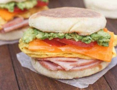 A Cheesy Egg, Avocado and Ham Breakfast Sandwich on an english muffin on a wooden table