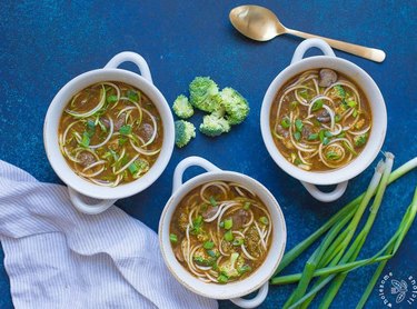 Instant Pot Spicy Beef and Broccoli Zoodle Soup in three white bowls topped with chopped scallions on a blue table.