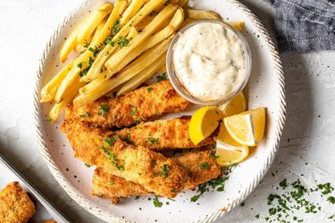 Air Fryer Fish and Chips on a white plate with fresh herbs, tartar sauce and lemon wedges
