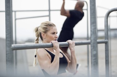 close up of a woman doing a chin-up exercise with a man doing chin-ups in the background