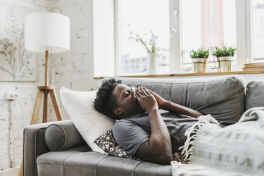 adult wearing a gray t-shirt lying down on a gray couch under a white blanket blowing their nose feeling dizzy from the flu