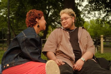 a LGBTQIA-representative couple sits outside looking at each other and smiling and supporting each other