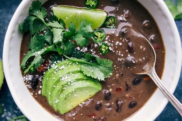 Close up of black bean soup in a white bowl topped with avocado lime and cilantro