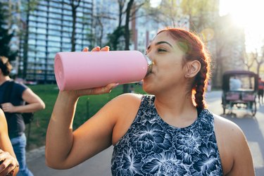 a young adult wearing a tank top and braids drinks electrolytes from a pink water bottle in a park outside