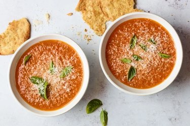 Two bowls of tomato soup on a white counter topped with basil leaves