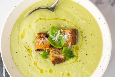 overhead photo of a bowl of light green homemade Cream of Broccoli soup. with croutons and a drizzle of olive oil