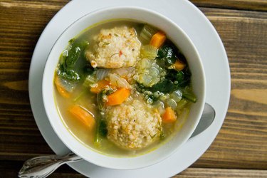 An overhead photo of a white cup of Italian Wedding Soup with veggies and chicken sausage on a wooden table