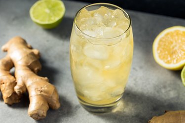 a glass of ginger ale with ice on a gray table, with pieces of ginger, lemon, and lime in the background