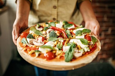 close view of a person holding a pizza topped with vegetables and vegan cheese, wondering if vegan cheese is healthy