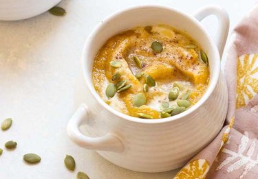 A small white cup of Vegan Autumn Glow Soup next to two spoons and scattered pumpkin seeds