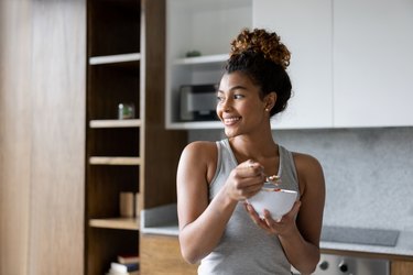 A woman stands in a kitchen smiling and eating from a bowl.